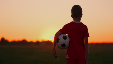 A-boy-standing-in-the-field-at-sunset-with-a-football-looks-into-the-distance-and-dreams-of-becoming-a-successful-football-player.-Stand-and-watch-the-sun-go-down-in-the-field